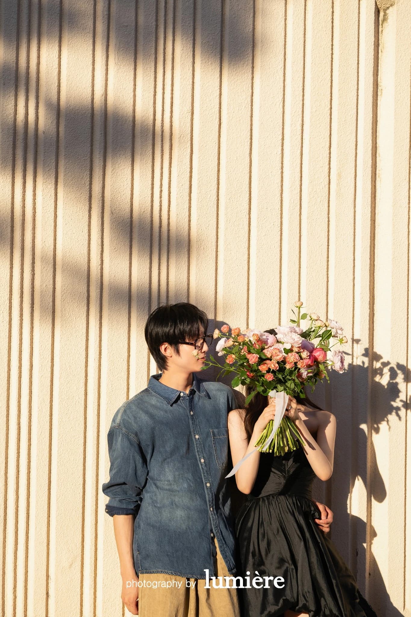 세로 홈이 있는 벽 앞 야외 공간에서 캐주얼 의상의 남녀 모델이 꽃다발로 얼굴 일부를 가리고 나란히 서 있음 모습이다. 꽃다발·데님 셔츠가 함께 보인다.