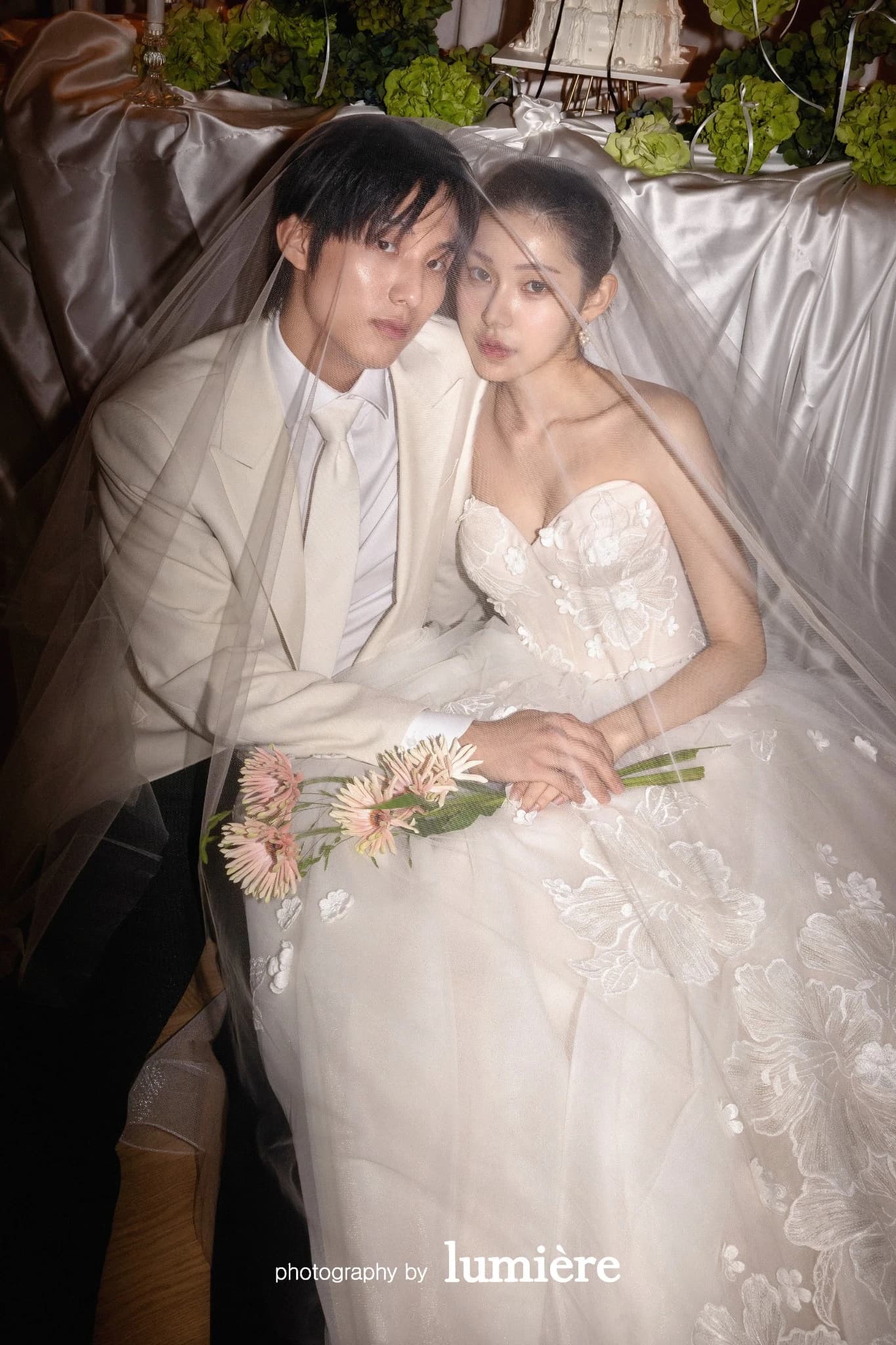 실내 테이블 장식 앞에서 웨딩 의상의 남녀 모델이 베일 아래에서 카메라를 바라봄 모습이다. 베일·부케가 함께 보인다.