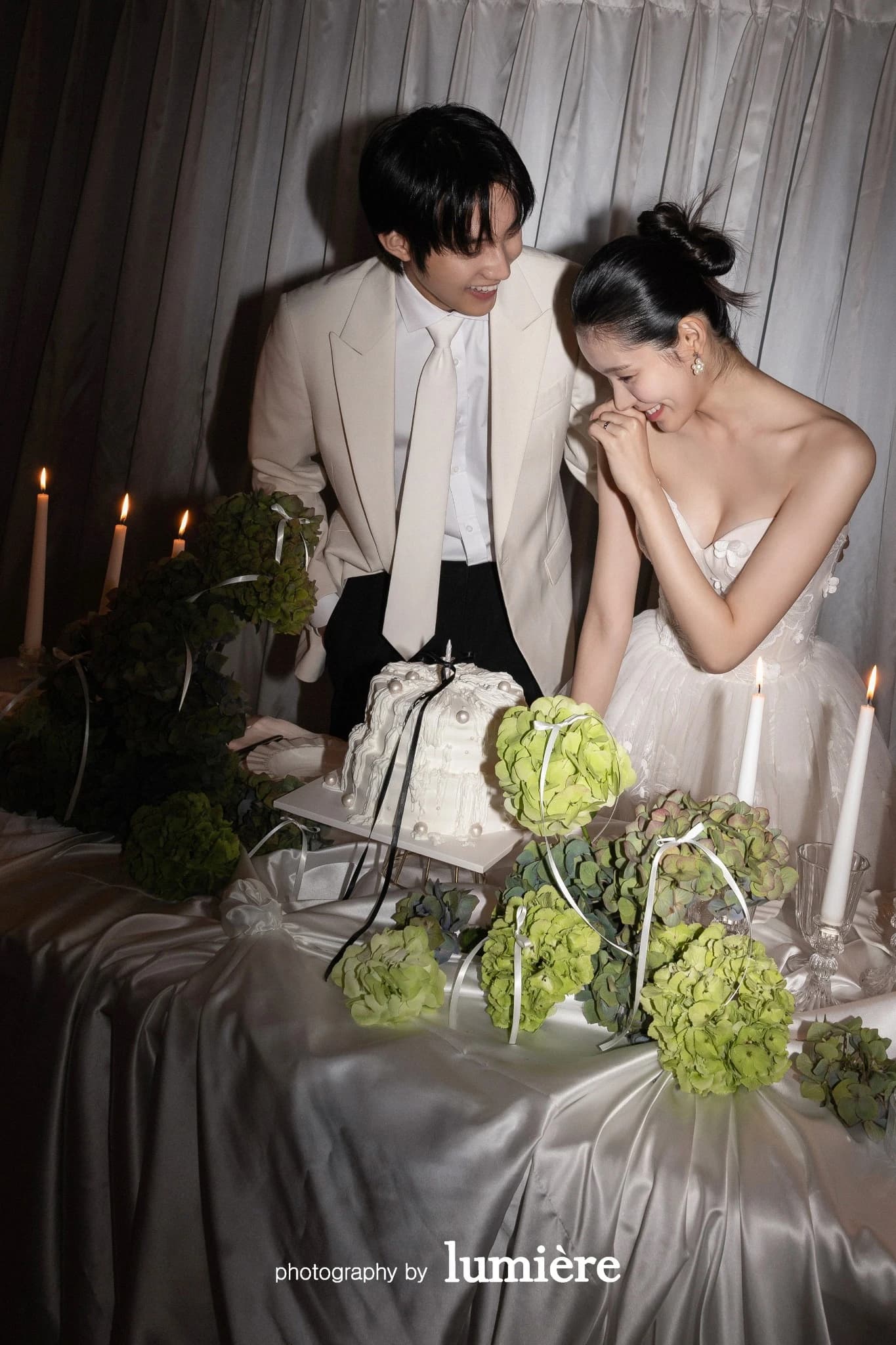 촛불과 케이크가 있는 실내 테이블 세트에서 웨딩 의상의 남녀 모델이 테이블 앞에서 웃는 표정을 보임 모습이다. 케이크·촛불이 함께 보인다.